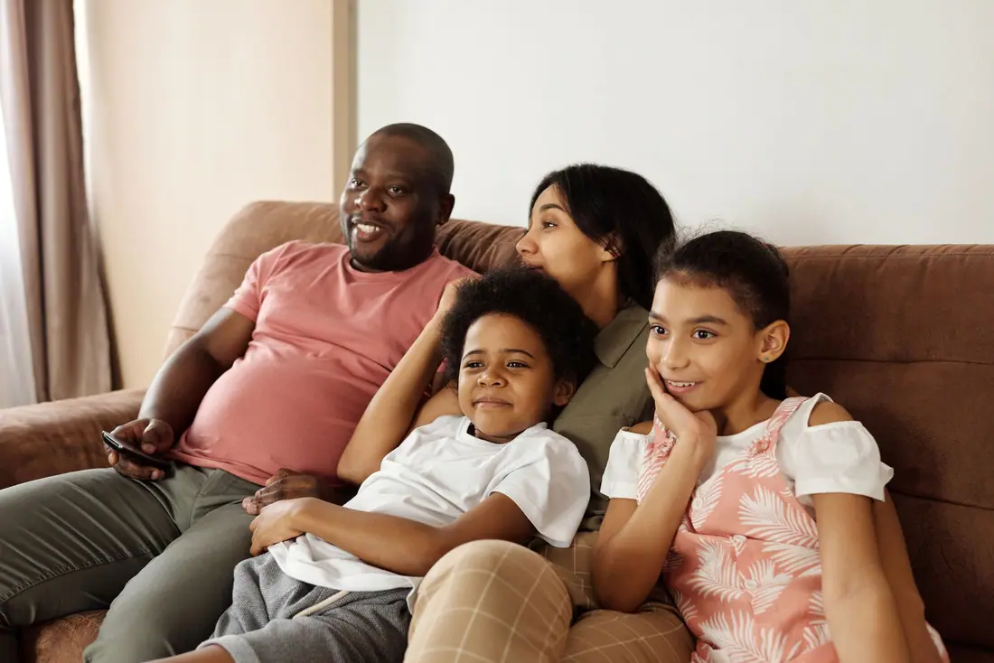 A smiling family of four leaning together on a brown sofa in a softly lit living room