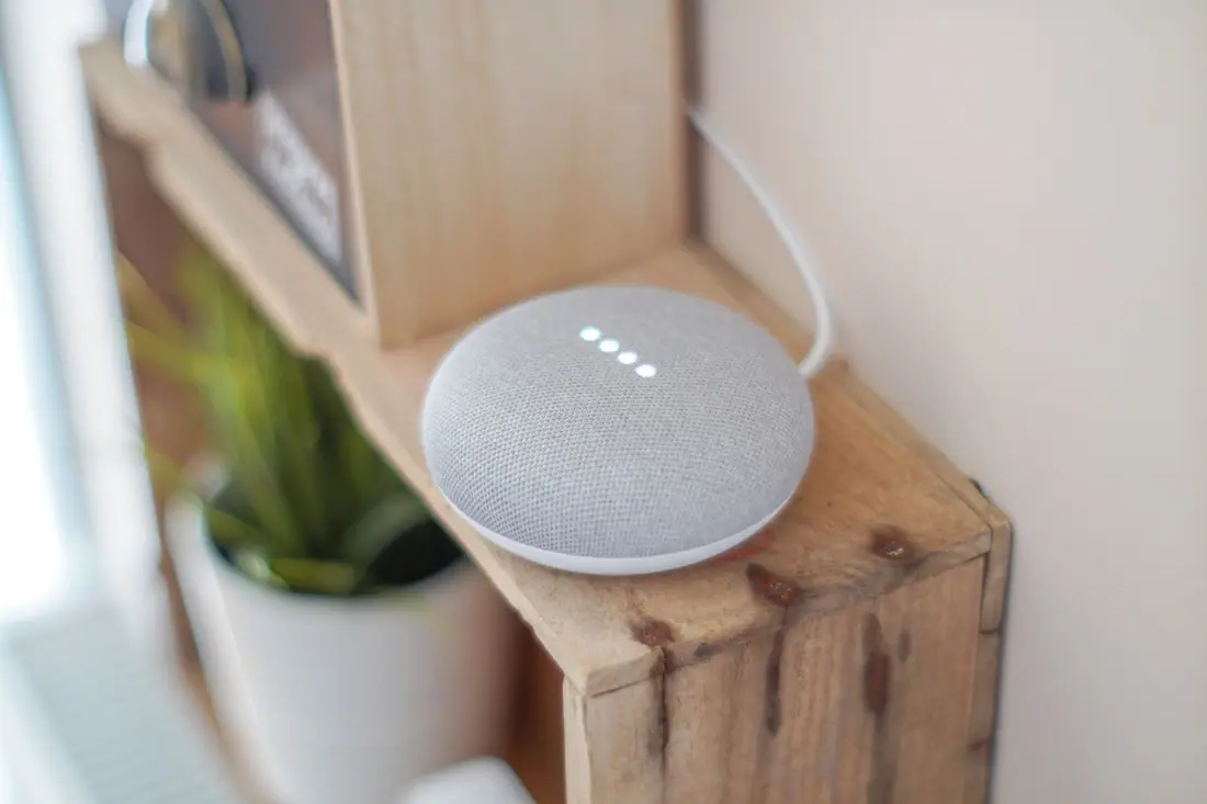 A small white fabric smart speaker on a wooden shelf beside a green plant in a bright room